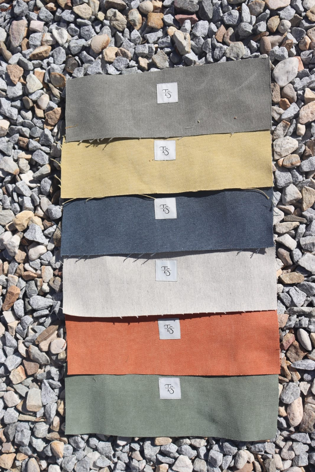 Six fabric swatches in different colors (olive green, burnt orange, grey, navy blue, mustard yellow, and olive green) arranged in a row on a bed of gray gravel, each with a small white label.