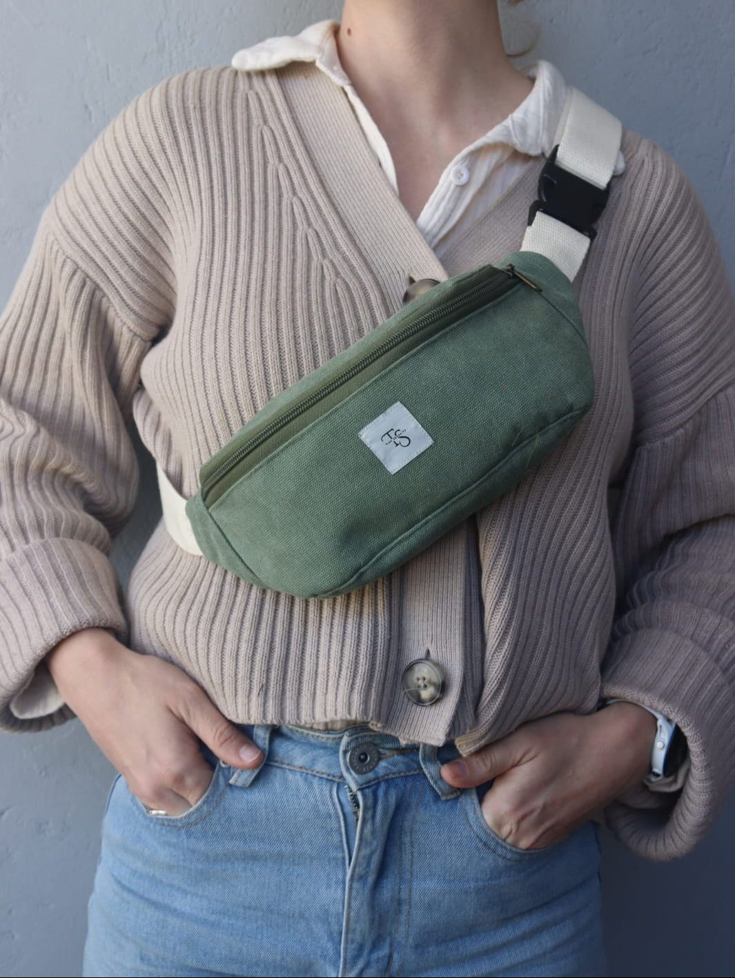A person wearing a beige ribbed cardigan and blue jeans carries an moss green canvas crossbody fanny pack with a white strap and a small white brand patch.